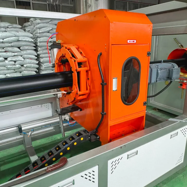 Orange downstream unit on a pipe extrusion line producing black plastic pipe in a factory setting with bags of material