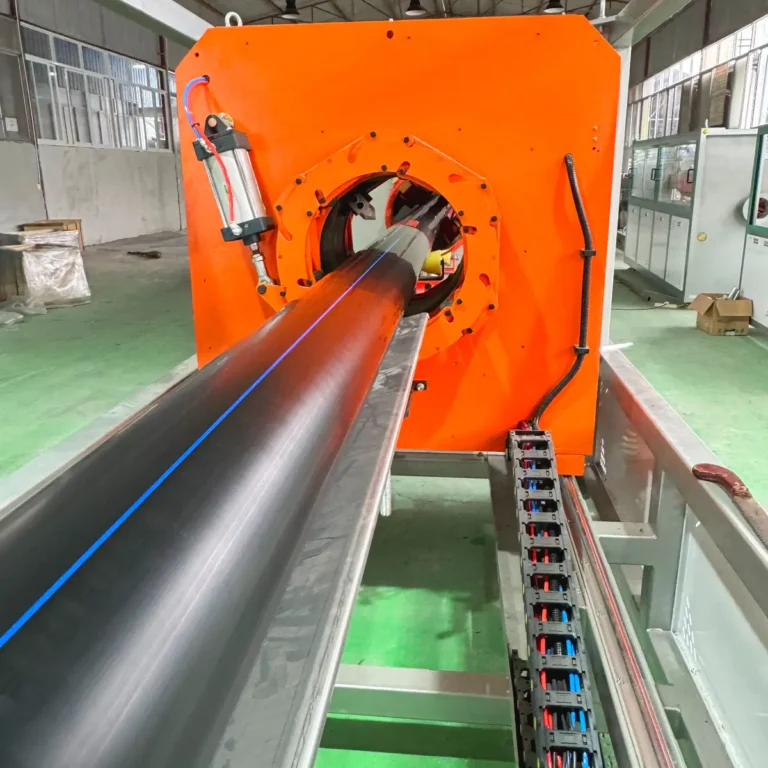 Large black plastic pipe exiting an orange extrusion unit on a factory floor with control cabinets in the background