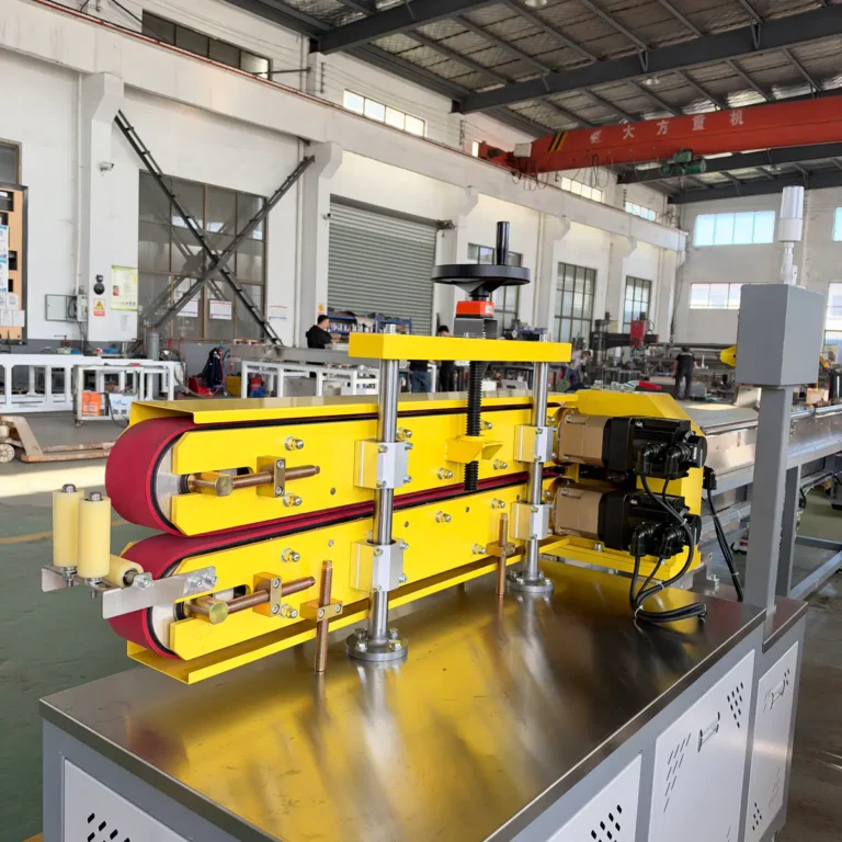 Yellow twin belt haul-off unit mounted on a metal base in a factory workshop with other extrusion equipment visible
