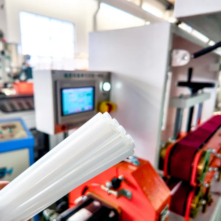 Hand holding several white plastic tubes in front of an extrusion machine and control panel on a factory floor