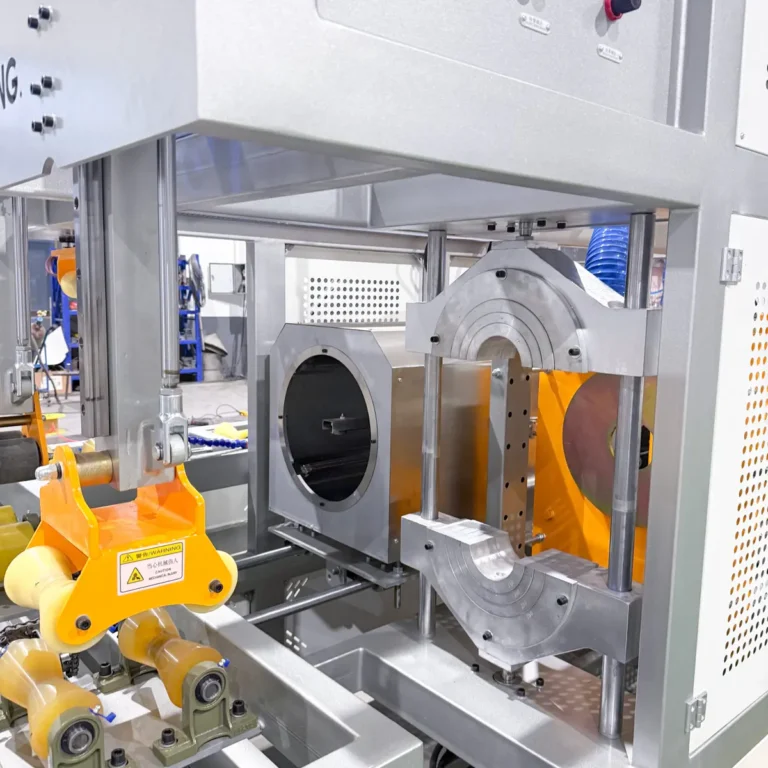 Closeup of a metal cutter and calibration section with yellow rollers on a PVC pipe extrusion line in a factory setting