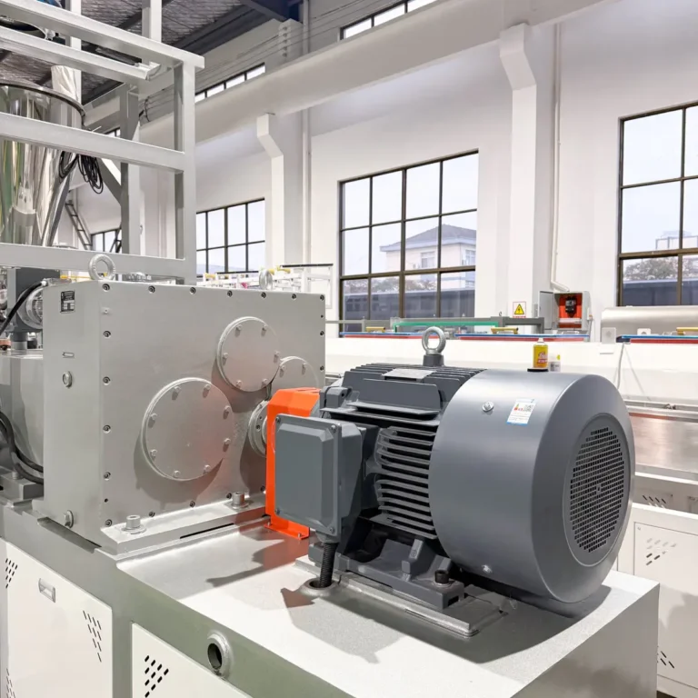 Extruder motor and gearbox assembly mounted on a machine base in a factory workshop with large windows in the background