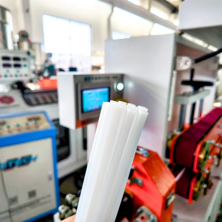 White plastic profiles are held in front of extrusion machinery and a control panel on a factory floor